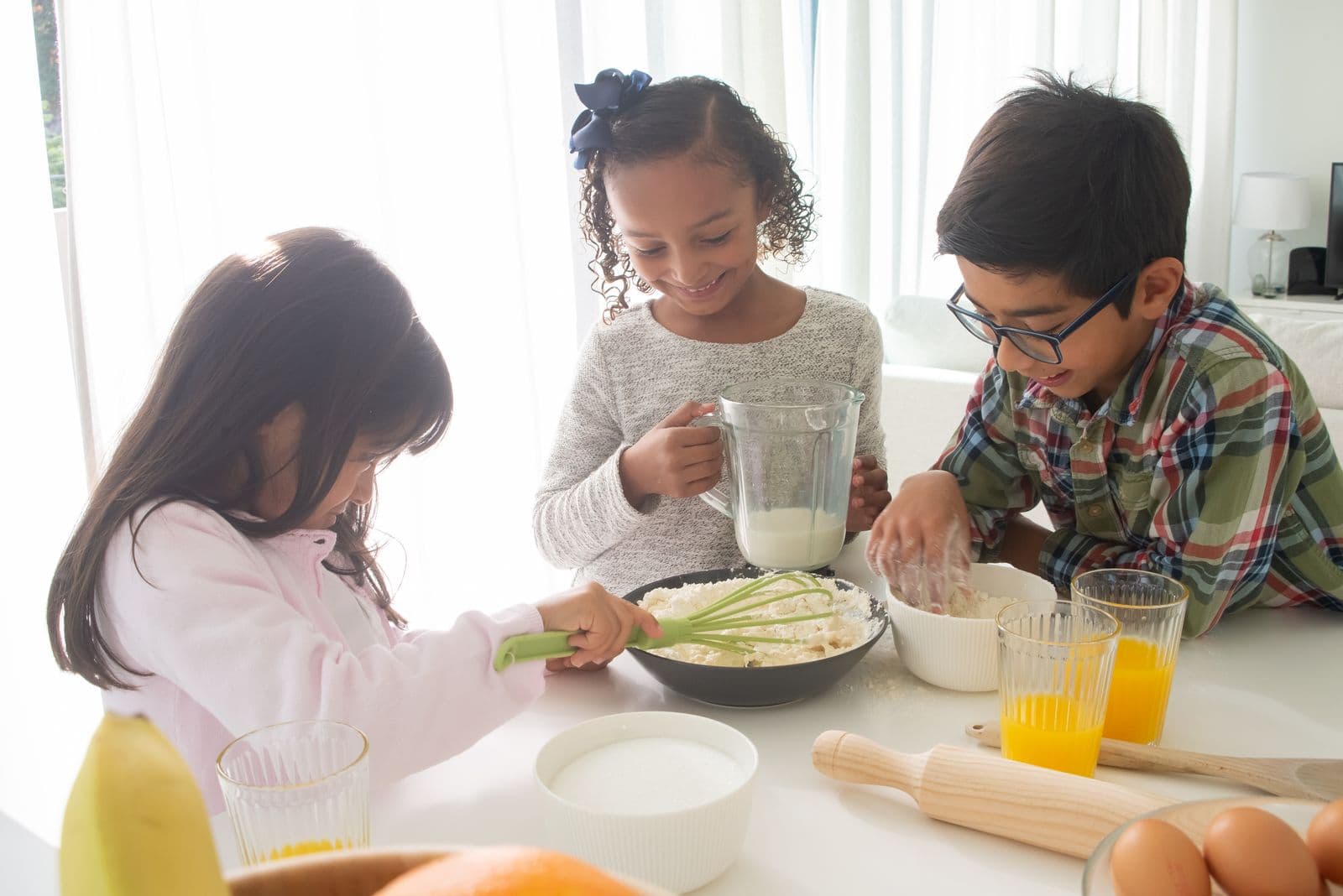 Trois enfants souriants préparent une pâte autour d'une table, avec un fouet, un pichet de lait, un rouleau à pâtisserie et des œufs.