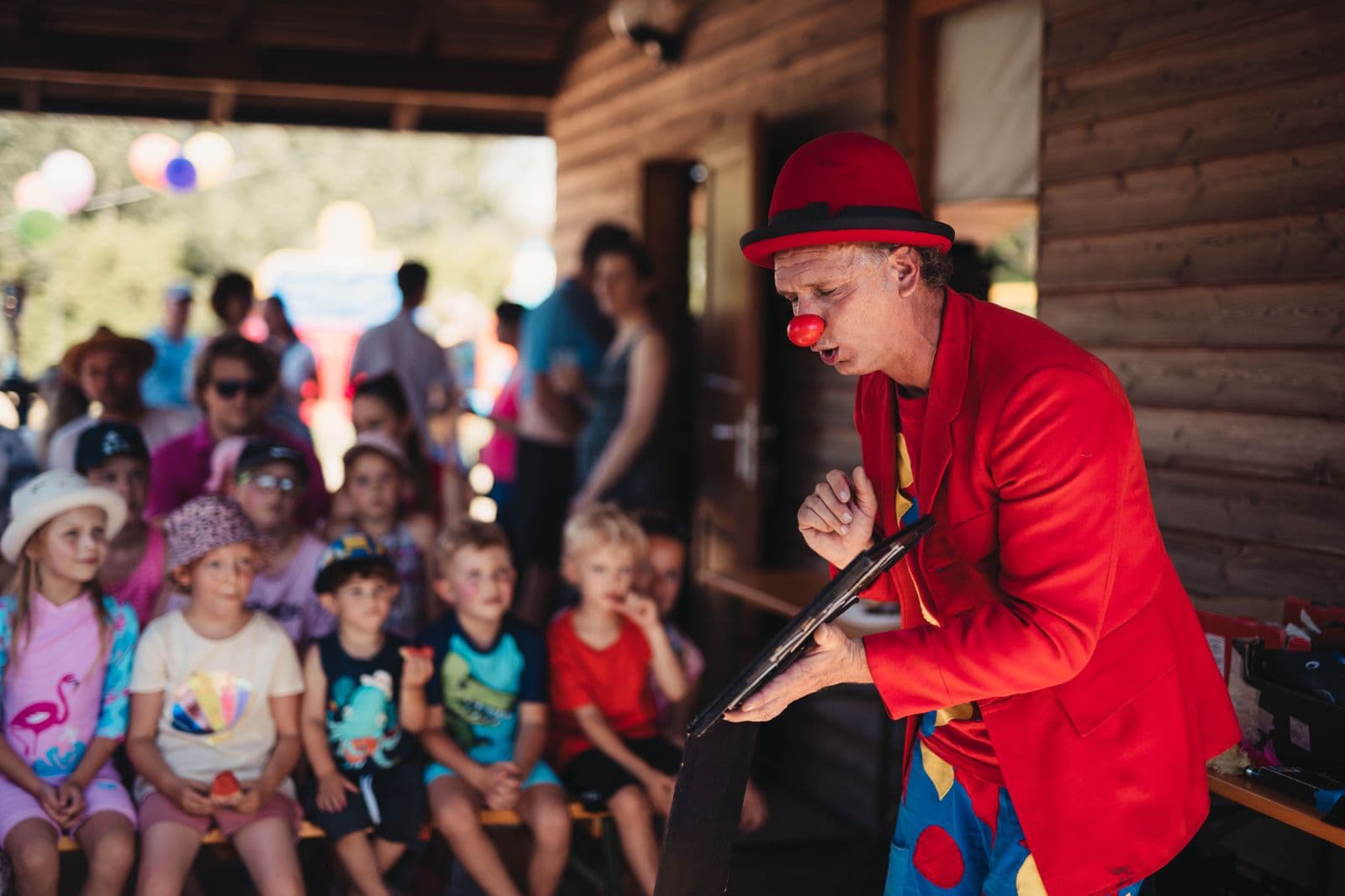 Un clown en veste rouge, chapeau rouge et nez rouge fait son numéro devant des enfants assis sur un banc sous un auvent en bois.
