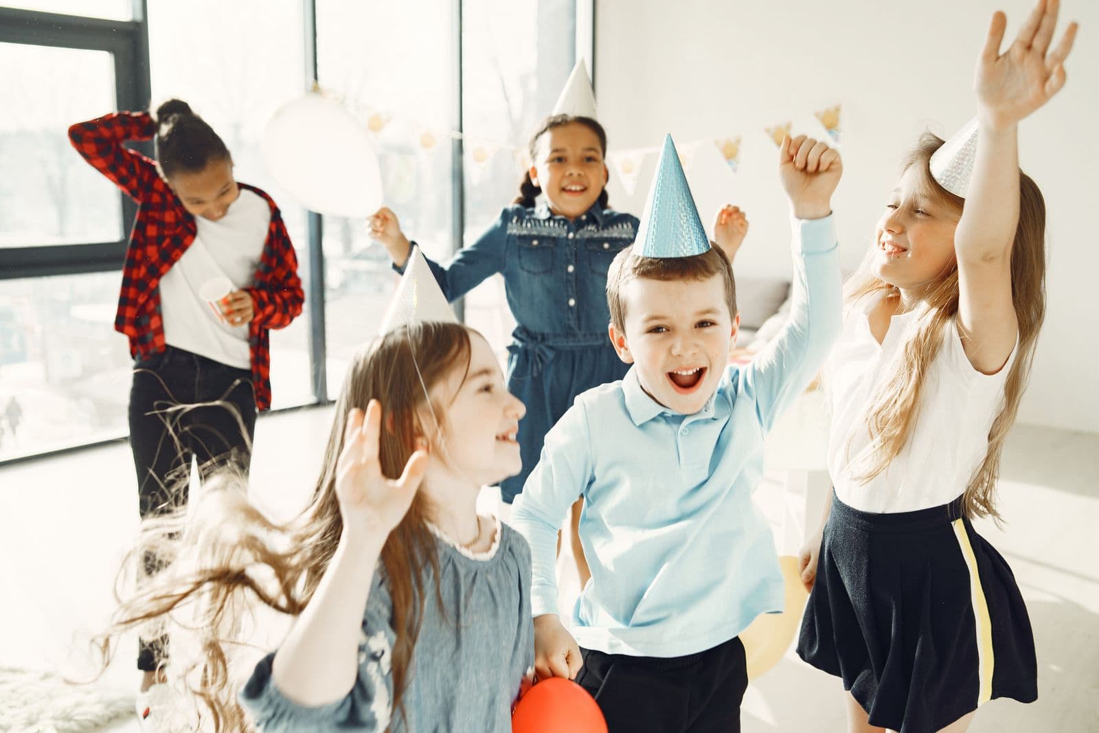 Des enfants coiffés de chapeaux pointus dansent et lèvent les bras dans une pièce décorée pour un anniversaire.