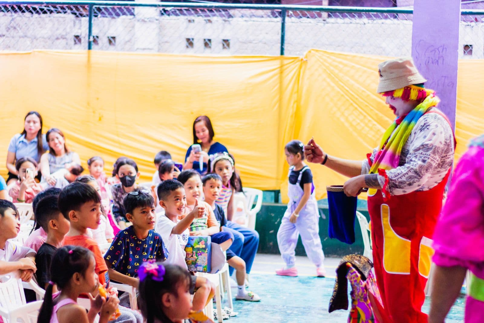 Un artiste en costume coloré présente un tour de magie avec un petit sac devant des enfants assis qui le regardent avec attention.