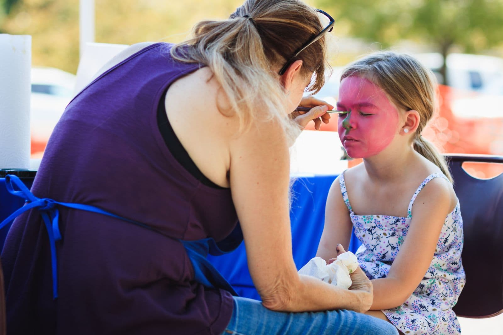 Une maquilleuse peint en rose le visage d'une fillette assise en plein air, les yeux fermés.