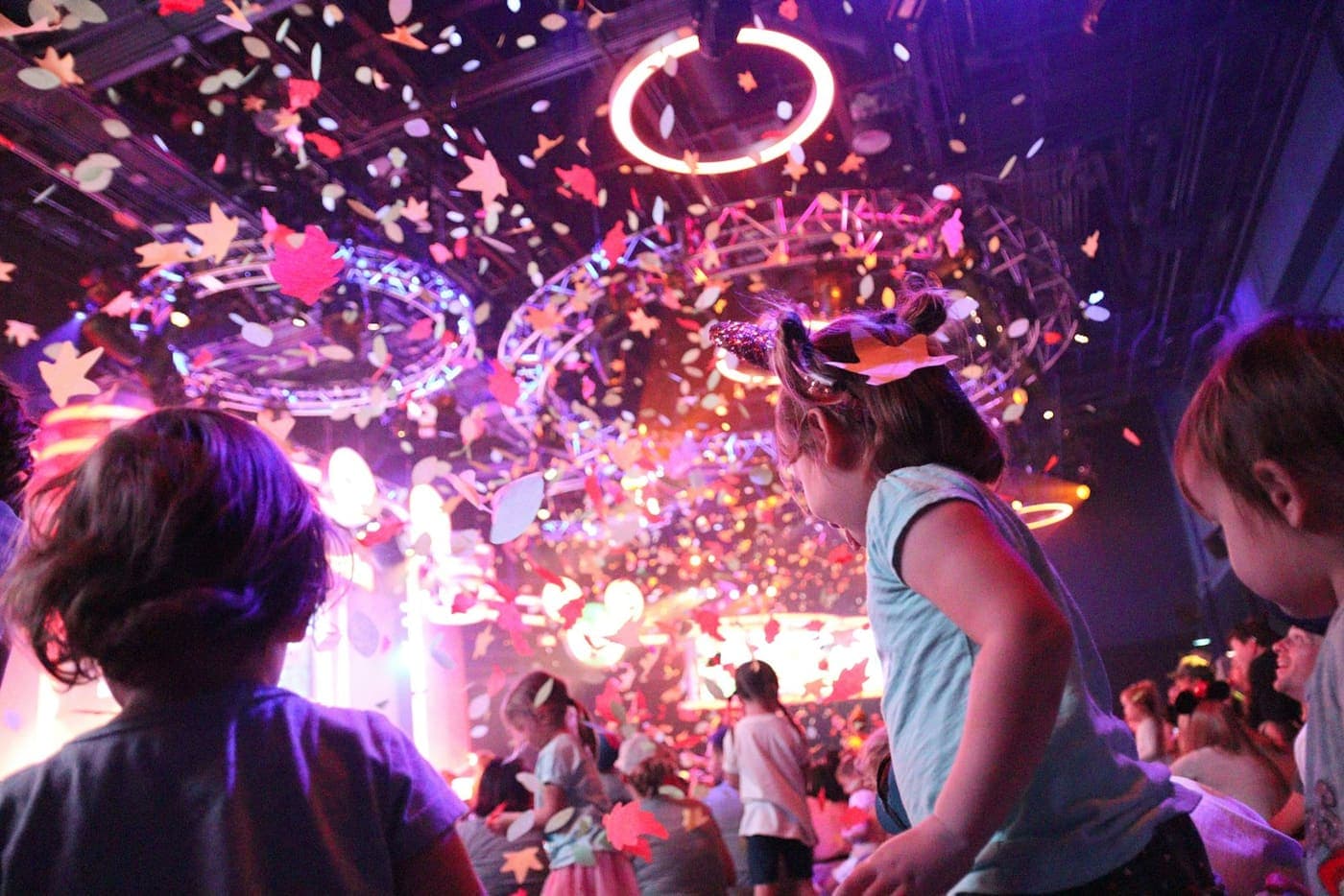 Des enfants regardent tomber des confettis dans une salle de spectacle éclairée en rose et violet