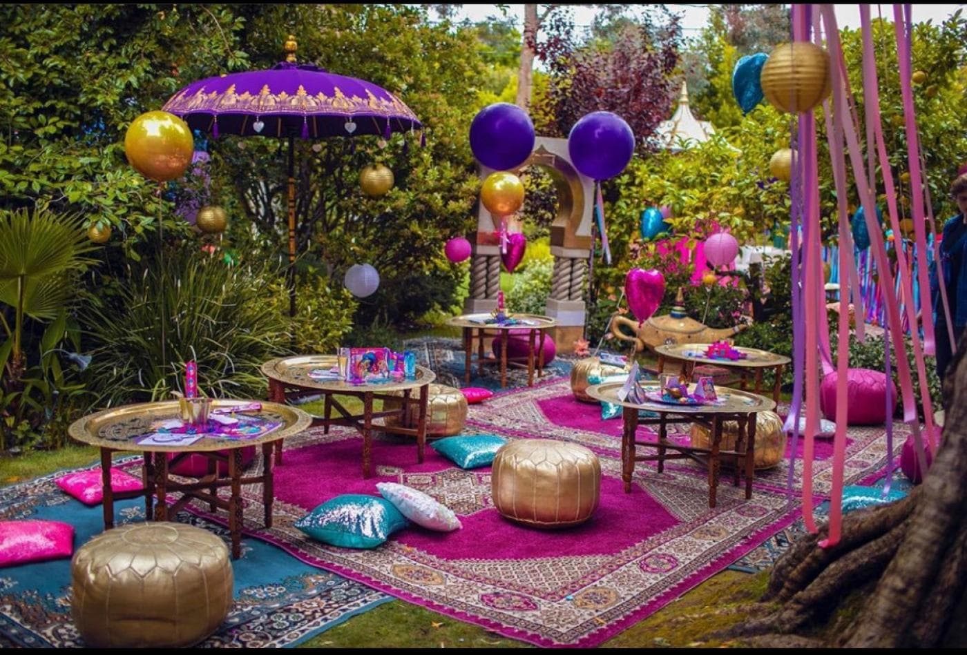 Fête d'anniversaire dans un jardin : tables basses en cuivre, poufs dorés, tapis, ballons violets et dorés